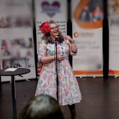 A woman in a floral dress with a flower in her hair speaks into a microphone.