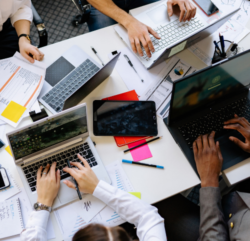 Aerial view of hands typing on laptops surrounded by notes and stationery.