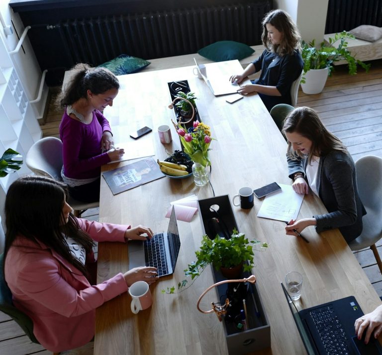 A group of five people working around a large table with laptops and plants.