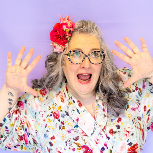 Vie in a floral dress and flowers in her hair, with her mouth open in surprise / shock and hands up by her face