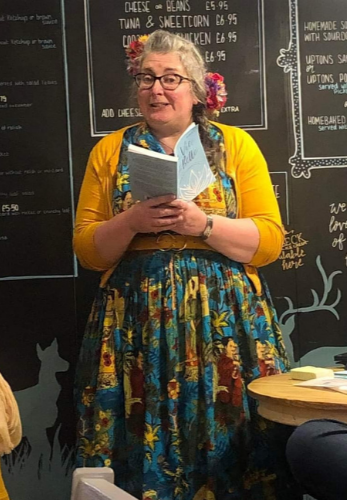 A woman in a colourful dress and yellow cardigan reads from a notebook.