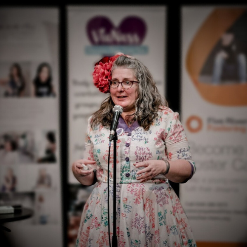 Vie in a floral dress with a flower in her hair speaks at a microphone.