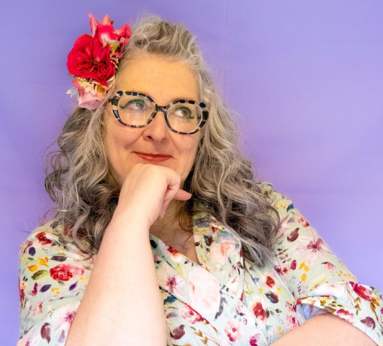 Vie with curly grey hair, glasses, and a floral blouse poses thoughtfully against a purple background.