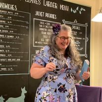 A smiling woman with curly hair and a floral dress stands in front of a menu board.