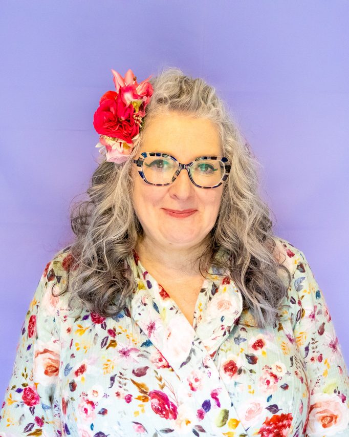 Vie with curly grey hair, flower accessory, and patterned shirt against a purple background.