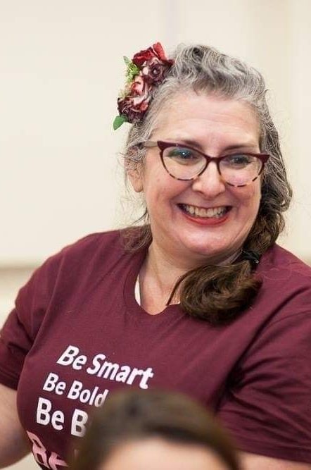 Smiling woman with glasses and floral hair accessory, wearing a maroon t-shirt.