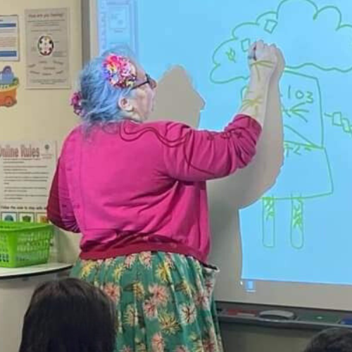 Vie draws a cartoon on a whiteboard while wearing a pink sweater.